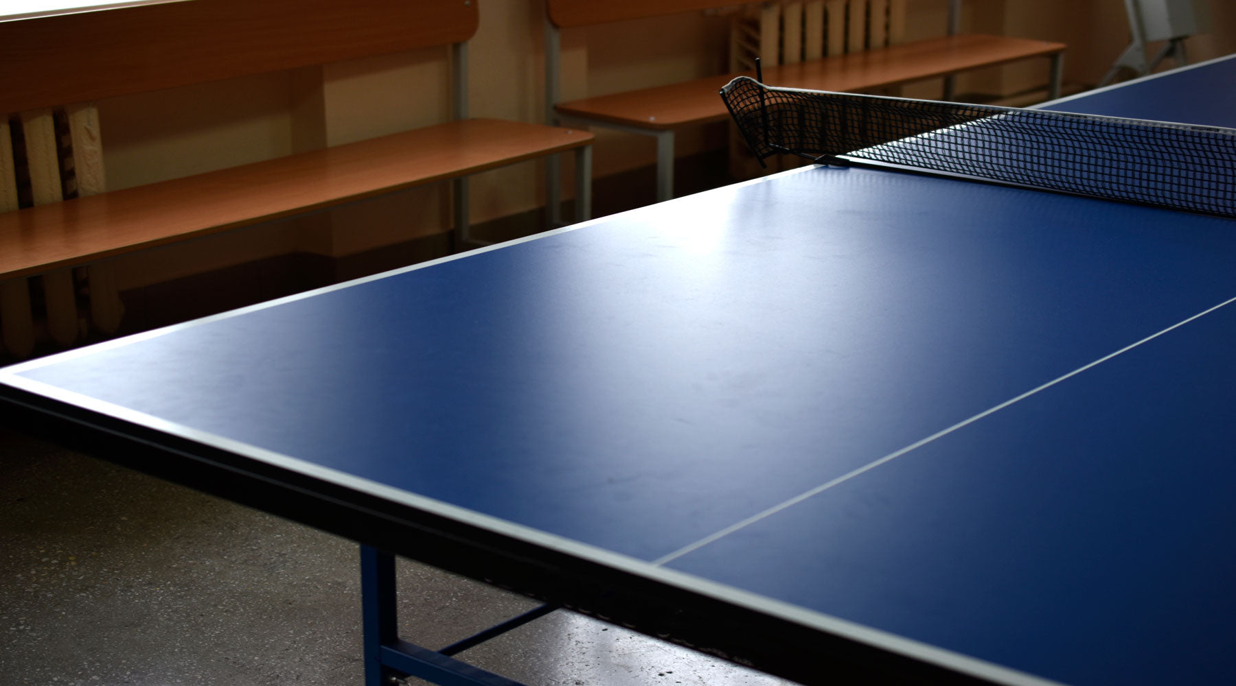 Indoor table tennis table with a blue playing surface and a net, set up in a room with wooden benches in the background